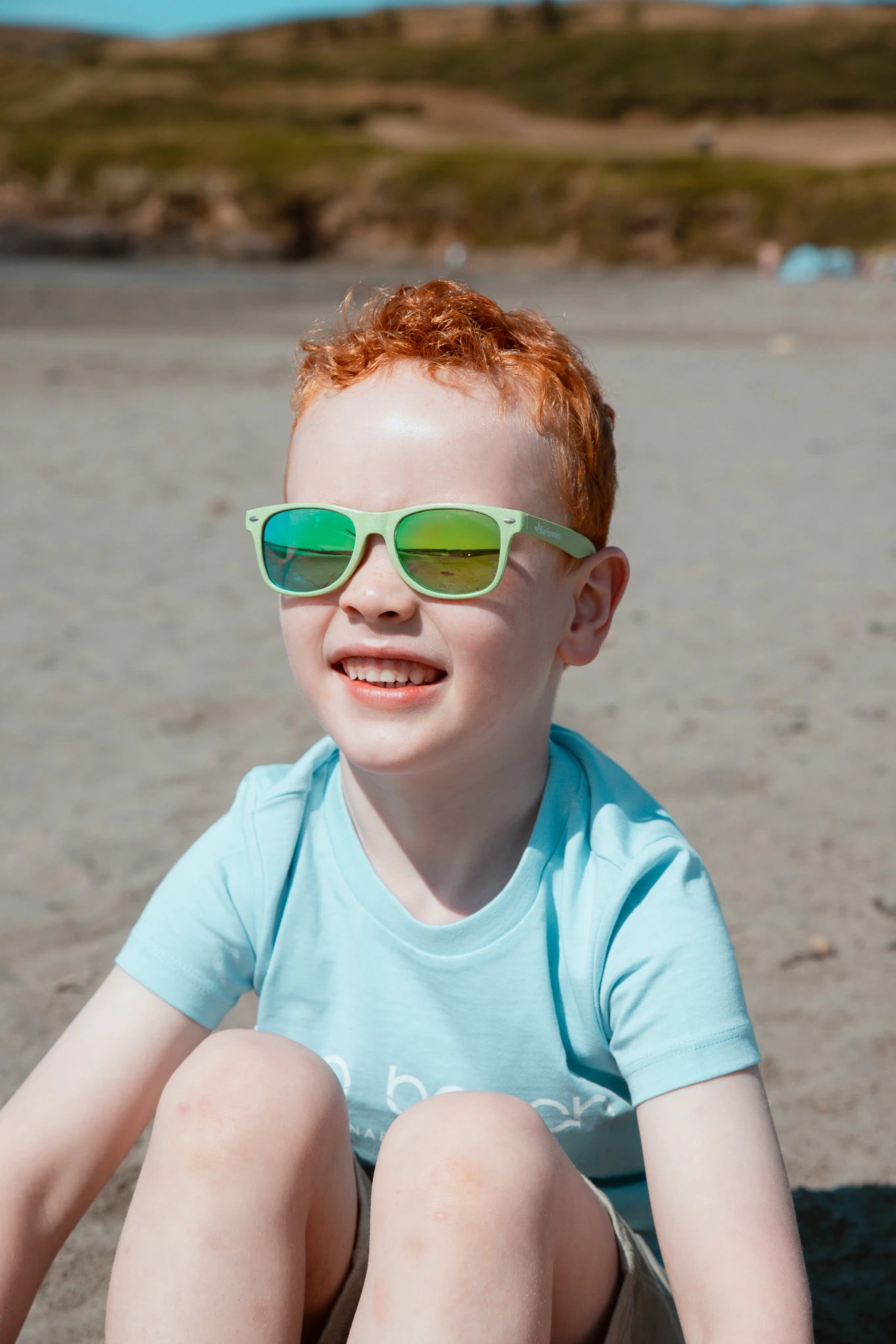 Child wearing green BioSunnies and a light blue shirt on a sandy beach.