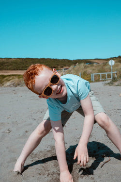 Child playing on a sandy beach with sunglasses and a blue shirt.