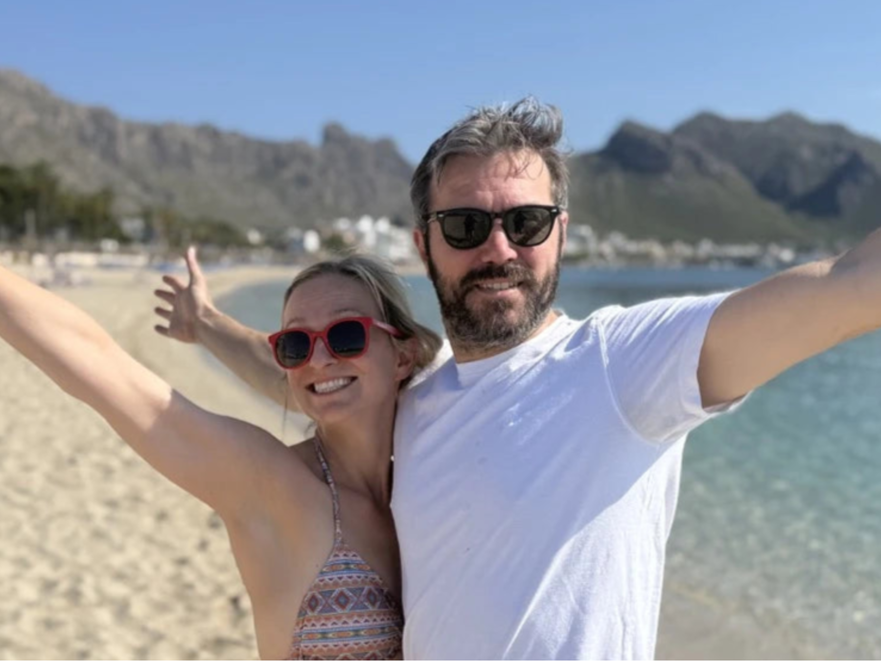 Two people standing on a beach with arms outstretched, mountains in the background