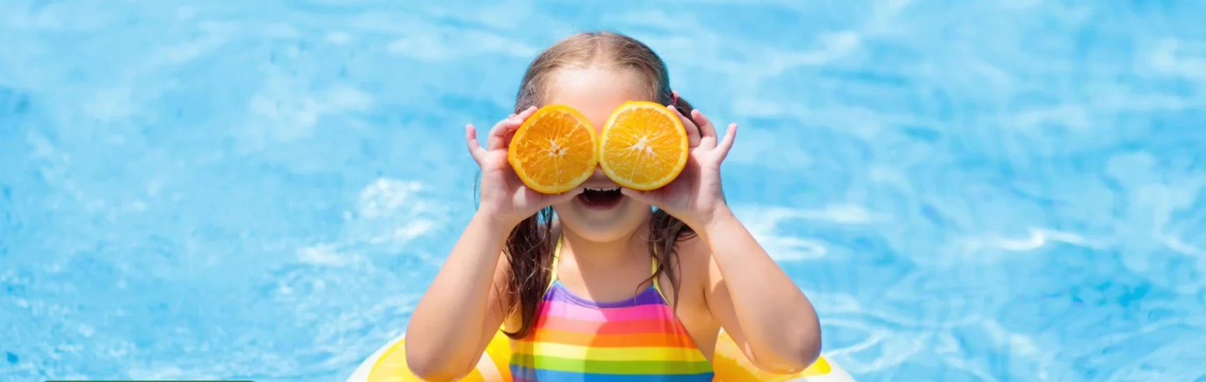 Child in a colorful swimsuit holding orange slices over their eyes in a pool.