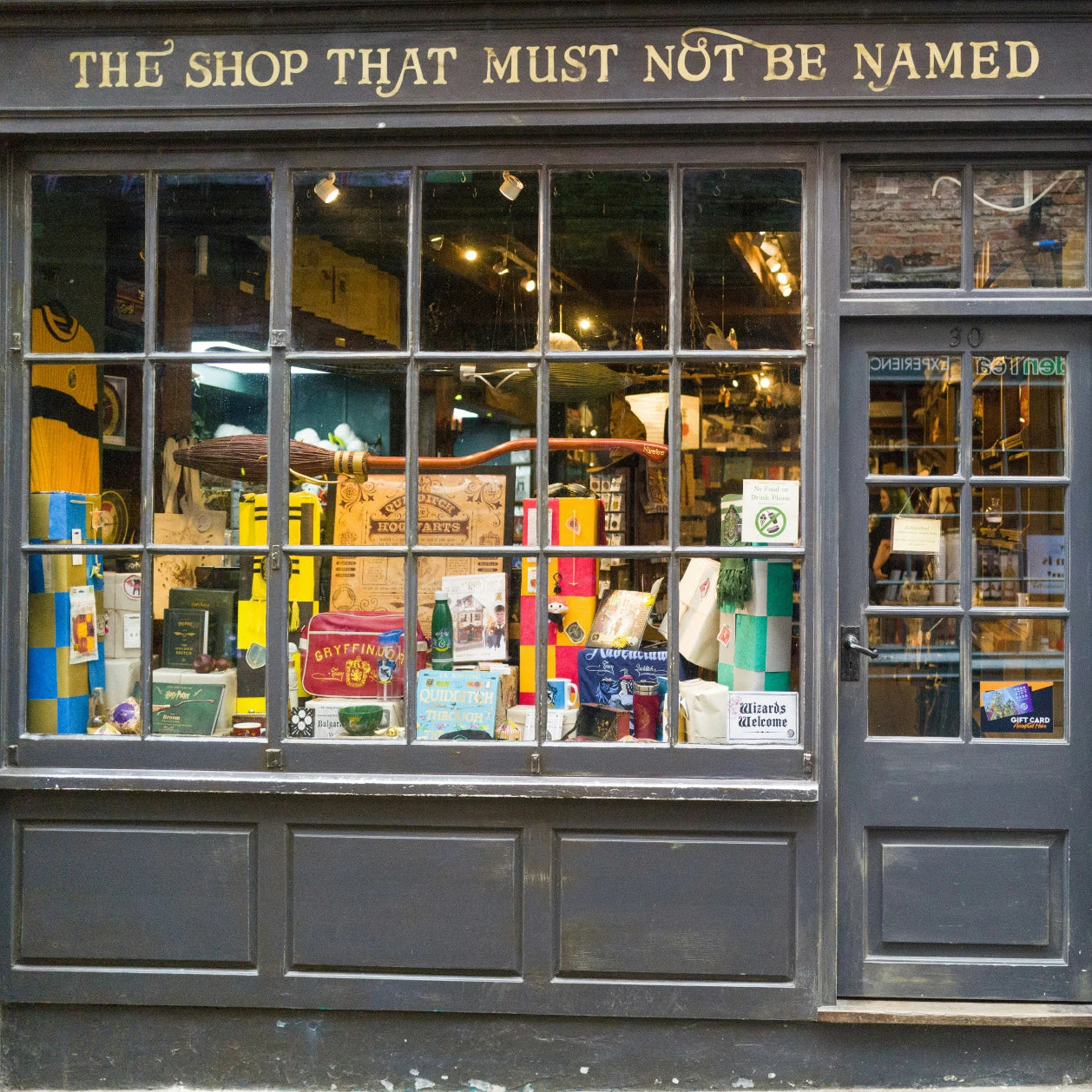 Shop front with glass window displaying various items and a sign above reading 'The Shop That Must Not Be Named'.